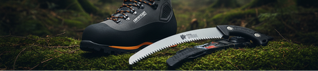 A black and gray outdoor hiking boot with orange accents lies on moss next to a folding camp saw with a silver blade.