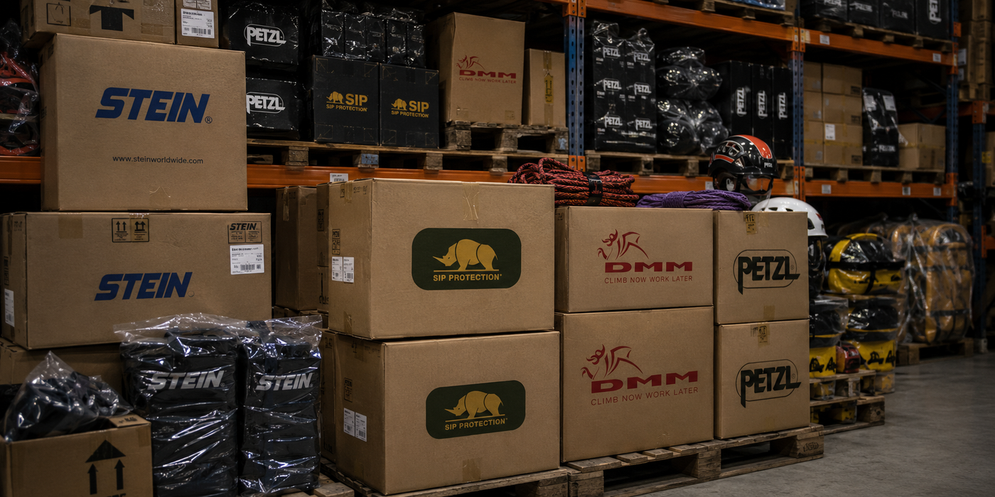 A warehouse or storage area with stacked cardboard boxes on pallets, including visible STEIN and PETZL branding. Shelves in the background hold more packaged items and equipment.